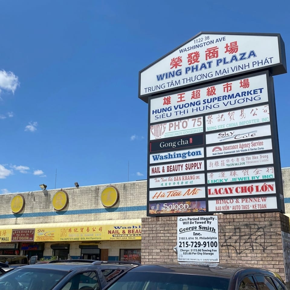 Wing Phat Plaza shopping center exterior with multilingual business directory, storefronts, parked cars, and blue sky.