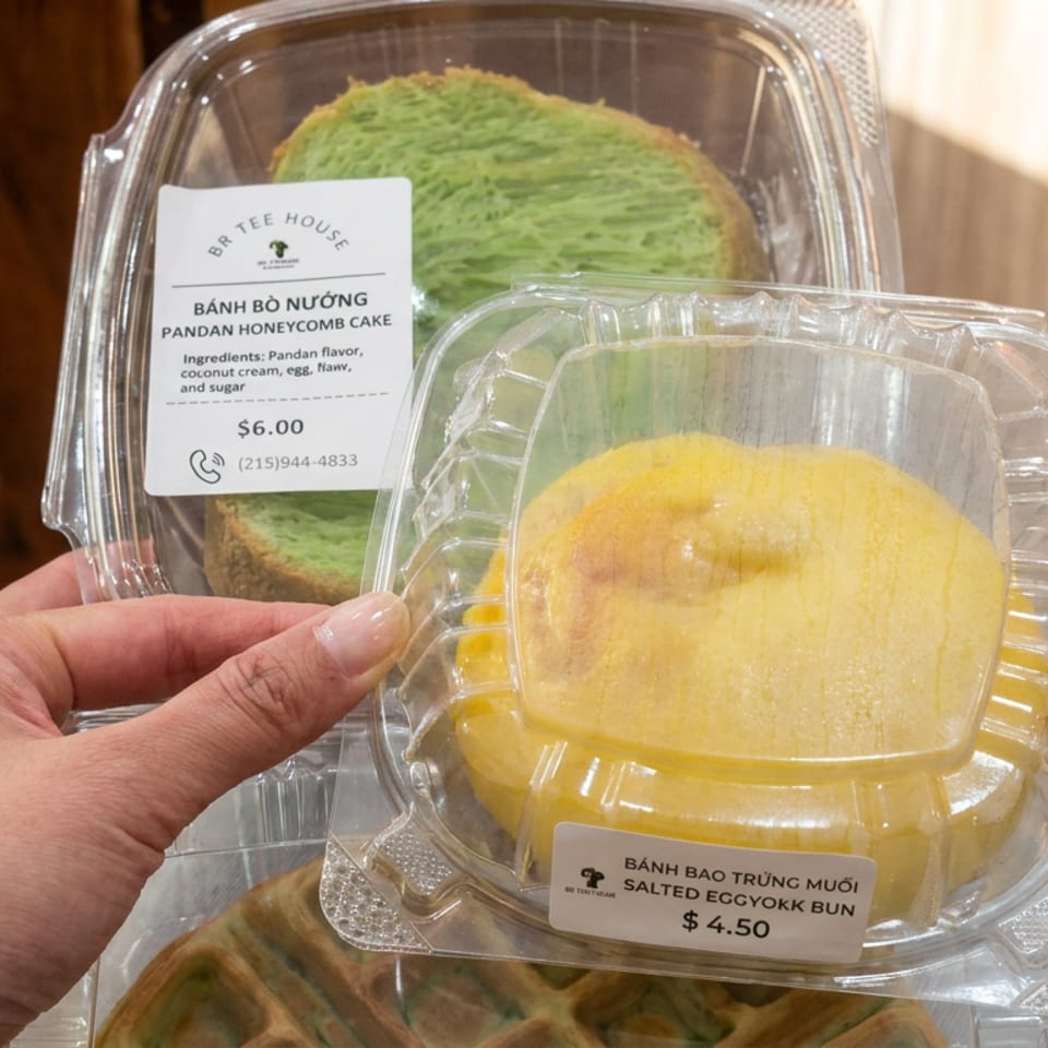 Hand holding packaged salted egg yolk bun, with pandan honeycomb cake and Vietnamese bakery labels visible behind it.