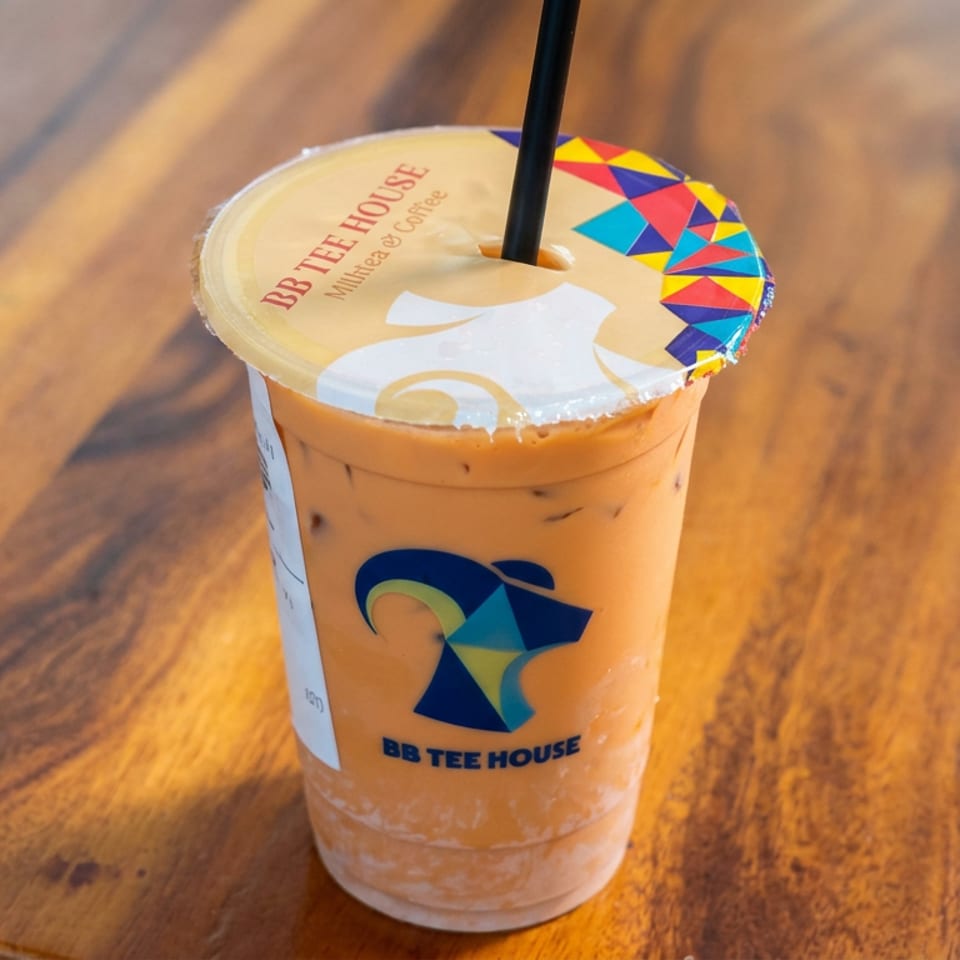 Iced milk tea in a BB Tee House cup with a colorful sealed lid and straw, resting on a wooden table.