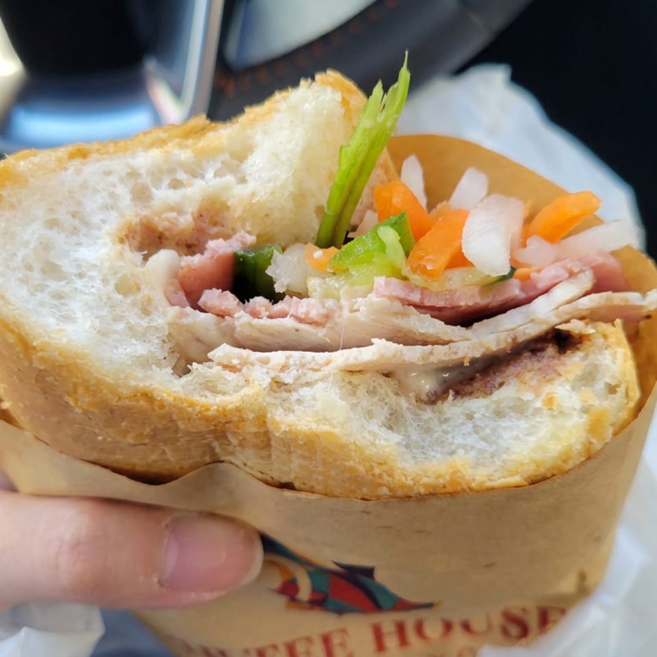 Hand holding a crusty baguette sandwich filled with sliced meat, chicken, pickled vegetables, and fresh herbs inside a car.