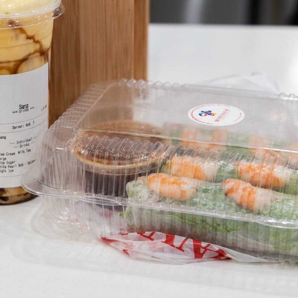 Packaged sushi rolls with shrimp and greens beside a taro bubble tea on a restaurant counter.