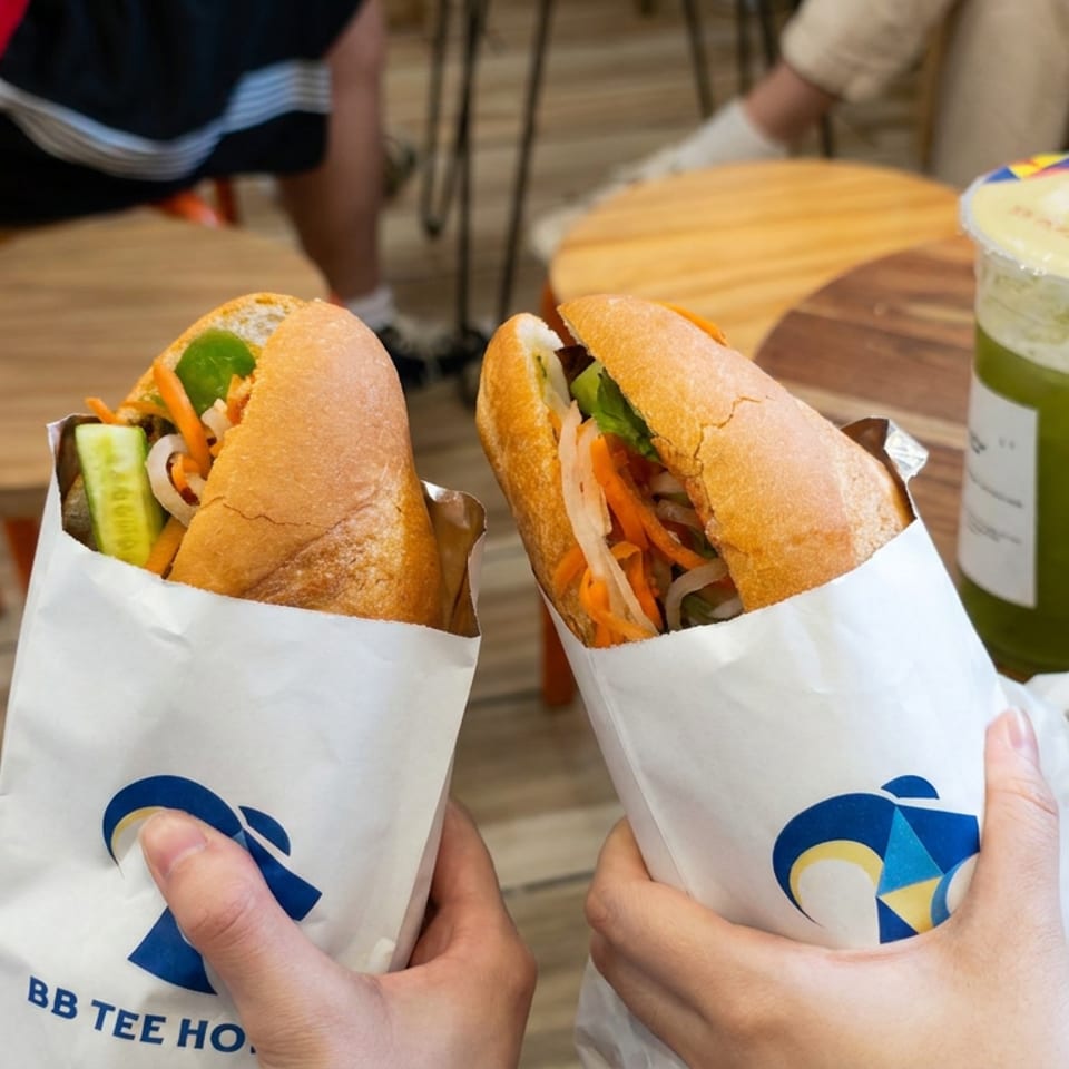 Two Vietnamese bánh mì sandwiches with pickled vegetables and cucumber held beside a green iced drink in a casual café.