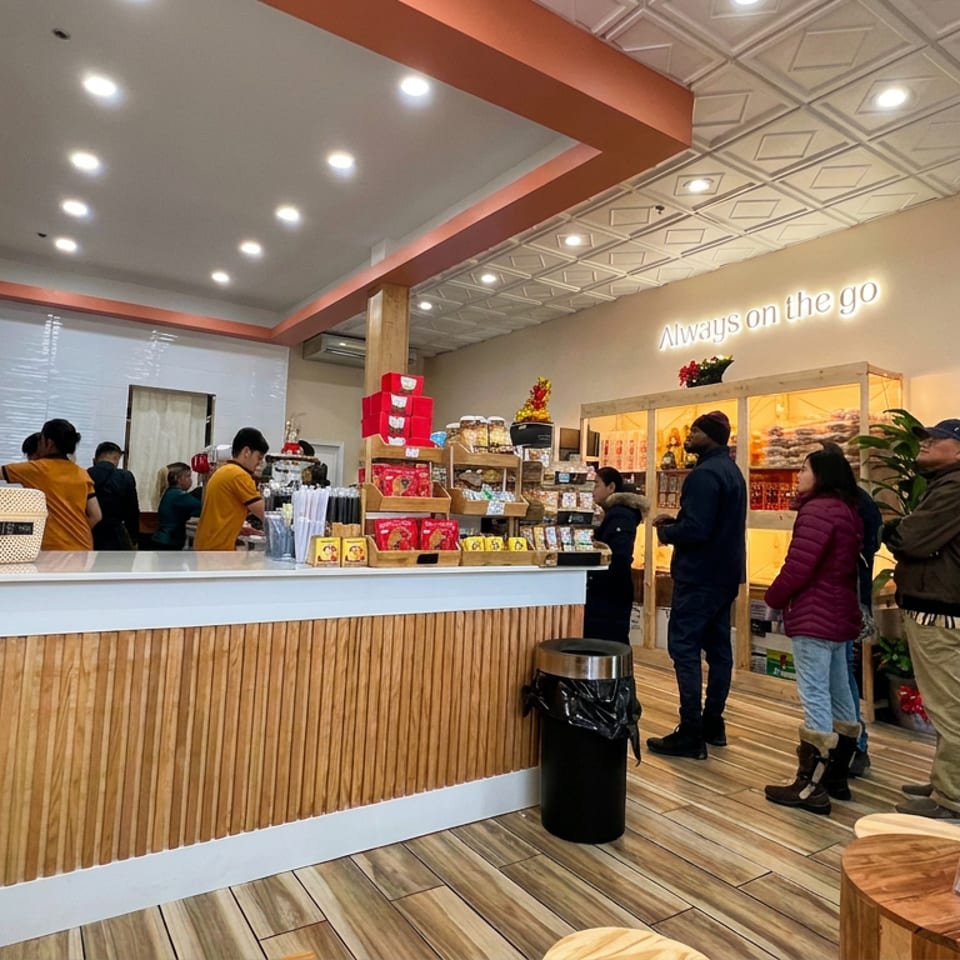 Busy bakery counter with staff serving customers, packaged treats on display, and warm wood-paneled decor beneath an “Always on the go” sign.