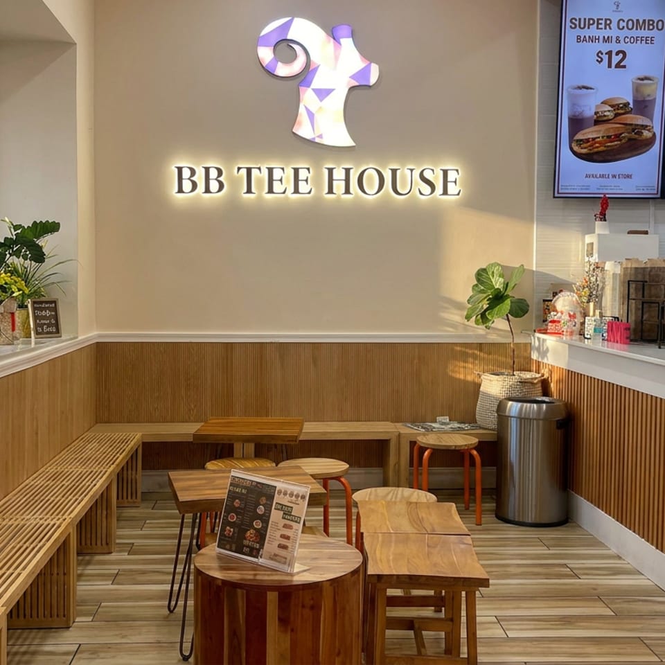 Warmly lit BB Tee House interior with wooden seating, counter service area, coffee equipment, plants, and illuminated logo.
