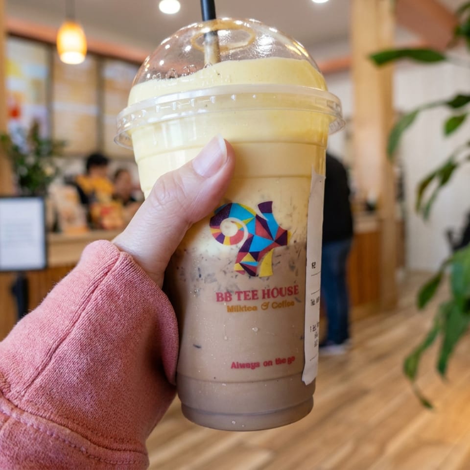 Hand holds a BB Tee House iced milk tea inside a warmly lit café with plants and patrons.